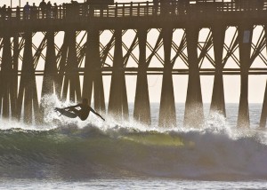 A surfer rides a wave with the Oceanside Pier in the background