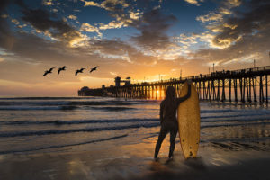 long hair surfer standing with surfboard at Oceanside Beach with Oceanside Pier at sunset